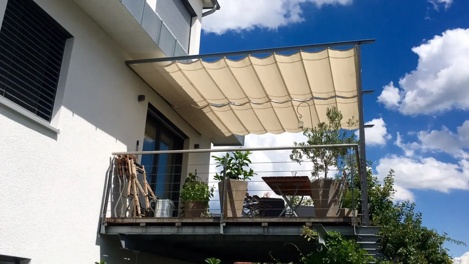 Balkon mit beiger Beschattung über dem Sitzplatz, davor Kübelpflanzen, Klapptisch und Stühle am Edelstahlgeländer